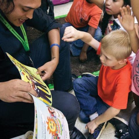 A volunteer reading a picture book to young children, several with their hands raised